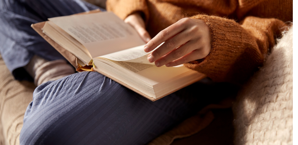 Eine gemütlich gekleidete Person sitzt im Schneidersitz, liest und blättert in einem aufgeschlagenen Buch.